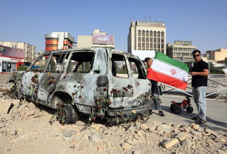 A man holds an Iranian flag by an Iranian Red Crescent ambulance that was destroyed during an Israeli strike, as seen here in Tehran on June 23, 2025. [Atta Kenare/AFP]